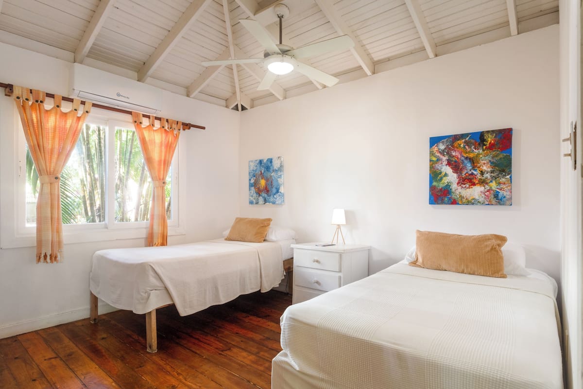 A room with two single beds, a ceiling fan, and two paintings on the walls.