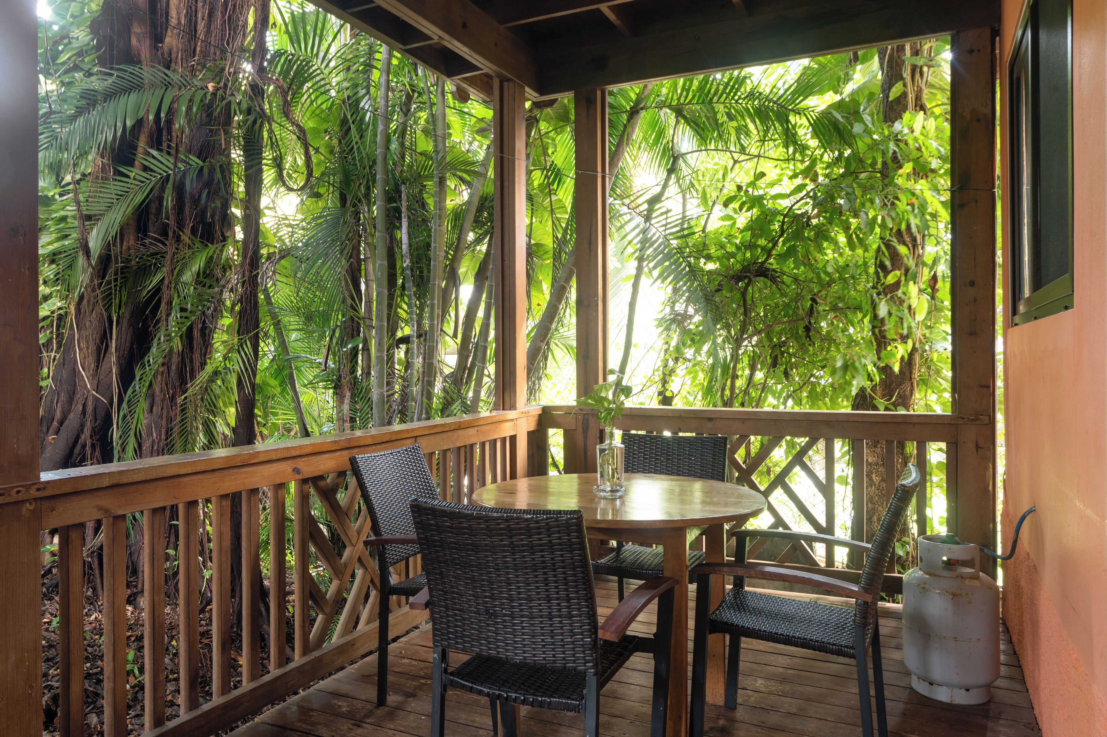A covered outdoor area with a wooden deck, wicker chairs, a round table, and a gas cylinder.