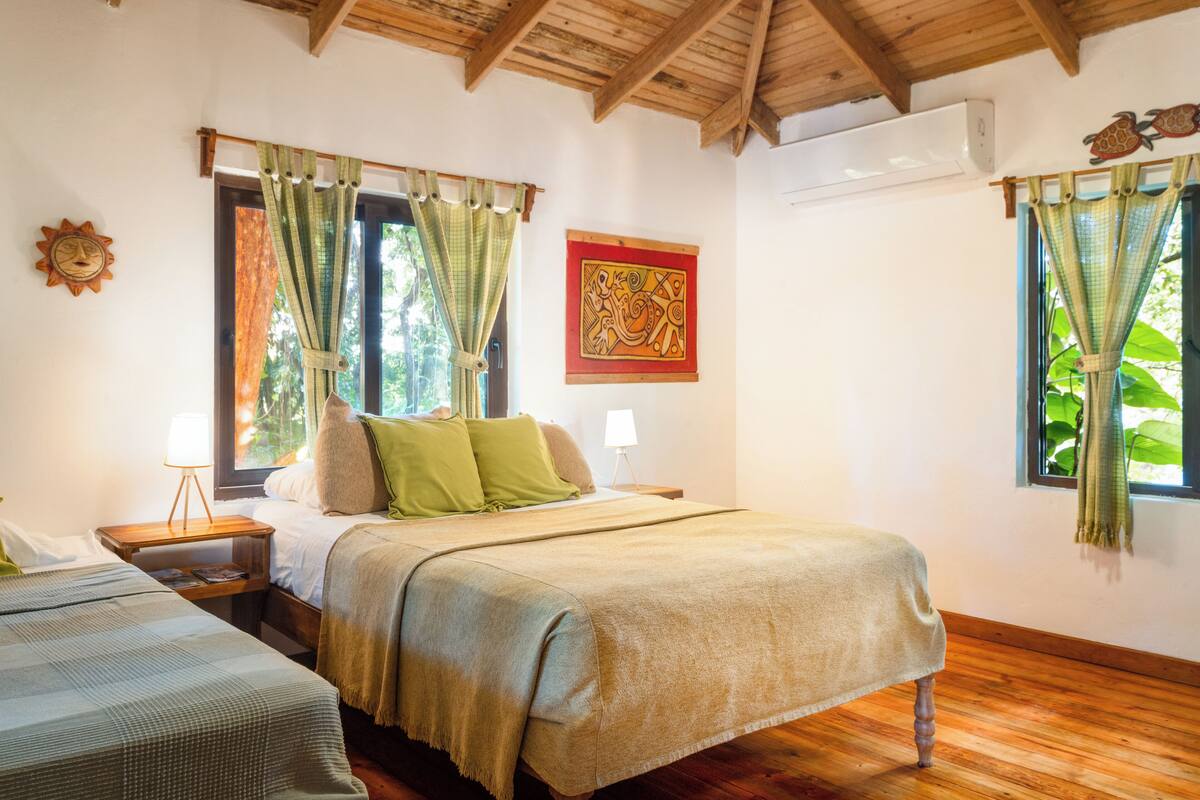 A bedroom with a bed, two bedside tables, a nightstand, a window with curtains, and a wall-mounted air conditioner.