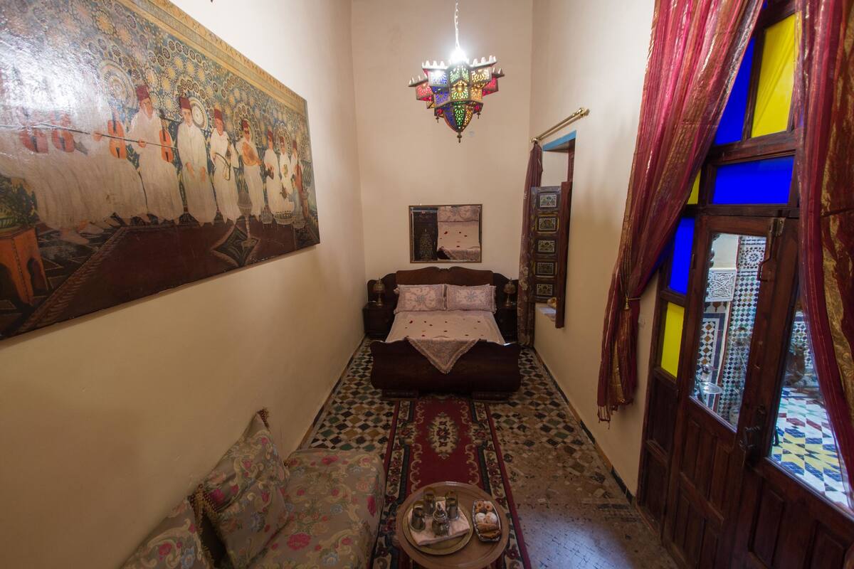 A narrow hallway with a bed, a chandelier, a large tapestry, and a patterned rug.