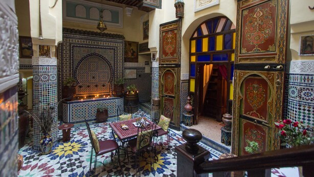 Un interior marroquí con suelo de mosaicos, una zona de estar central y paredes ornamentadas.