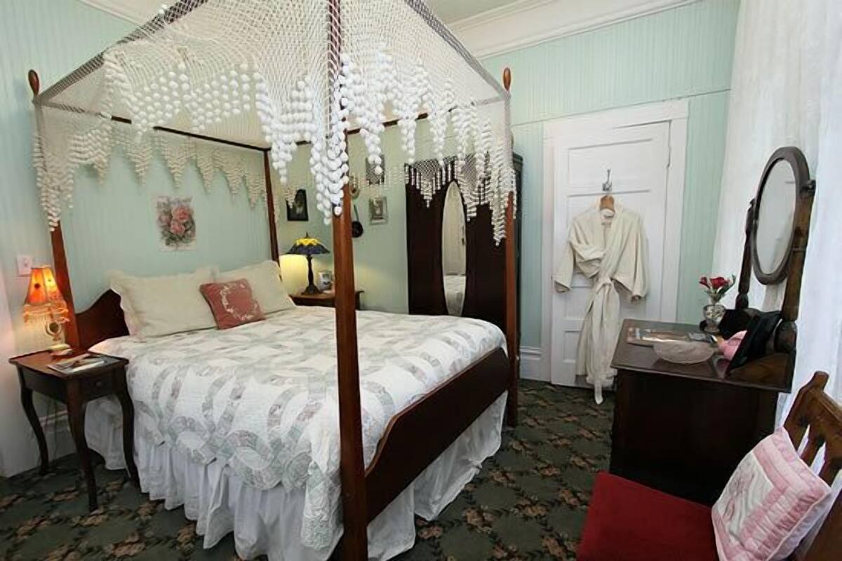 A four-poster bed with a canopy, a bedside table with a lamp, a dresser, a mirror, and a door with a robe hanging.