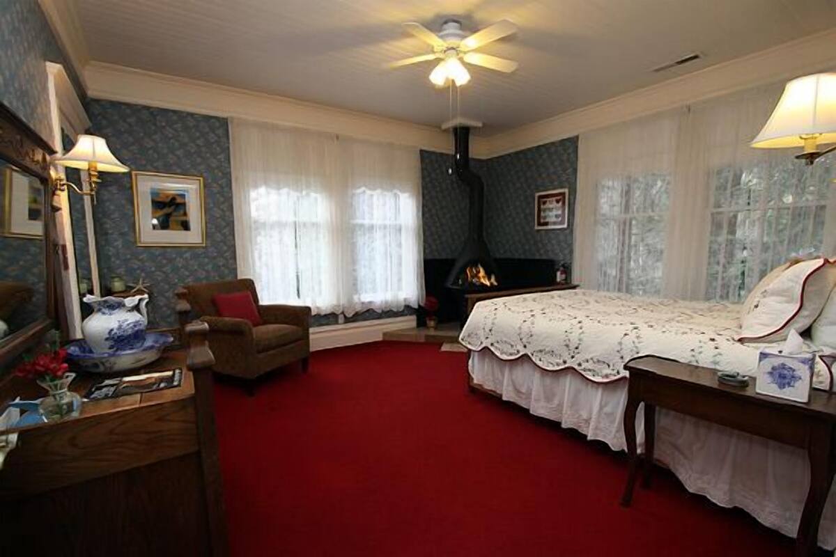 A bedroom with a red carpet, a bed with a white bedspread, a fireplace, a ceiling fan, and a desk with a lamp.