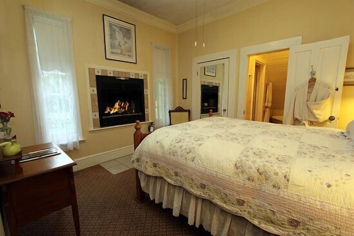 A bedroom with a fireplace, a bed with a floral bedspread, a wooden nightstand, and a mirror on the wall.