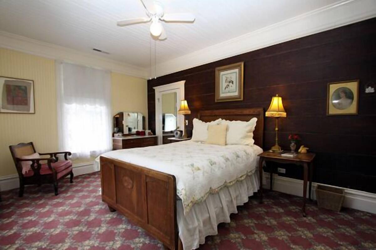 A bedroom with a wooden bed, a chair, a nightstand with a lamp, and a ceiling fan.