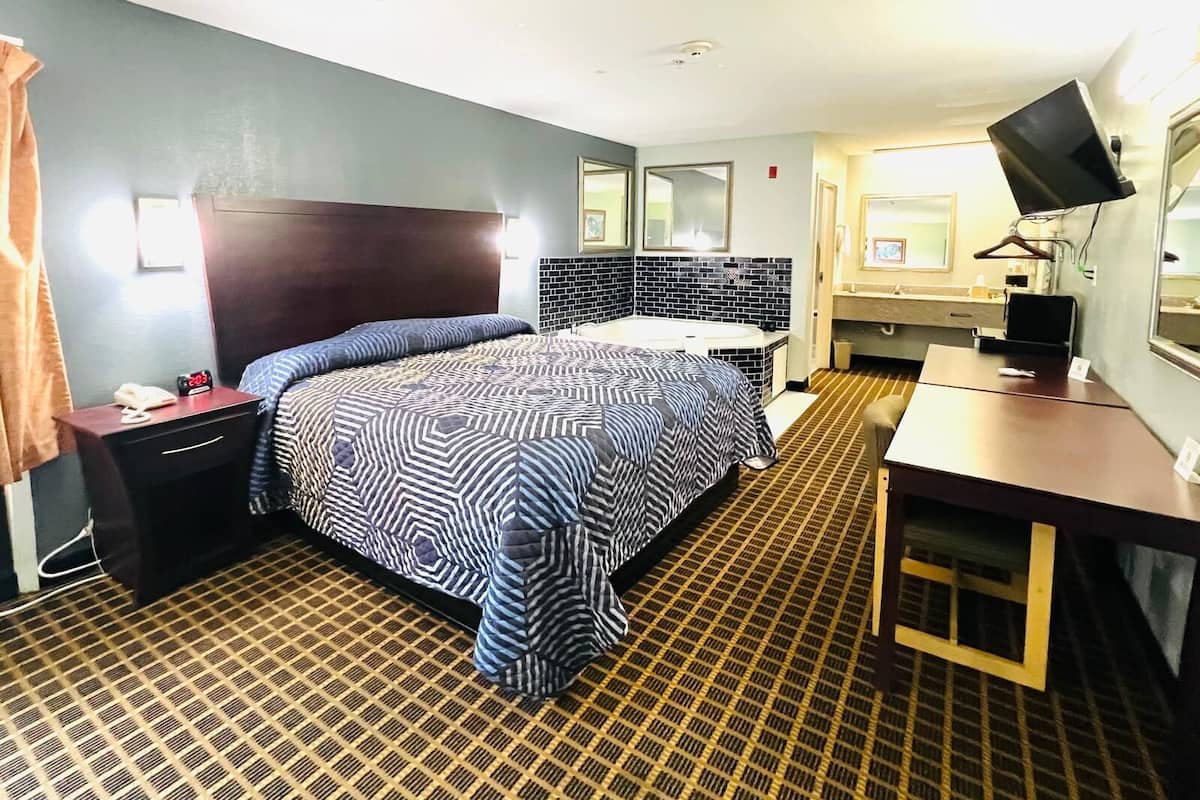 Quarto de hotel com cama, escrivaninha, cadeira, televisão, espelho, mesa de cabeceira e banheiro com pia e espelho.