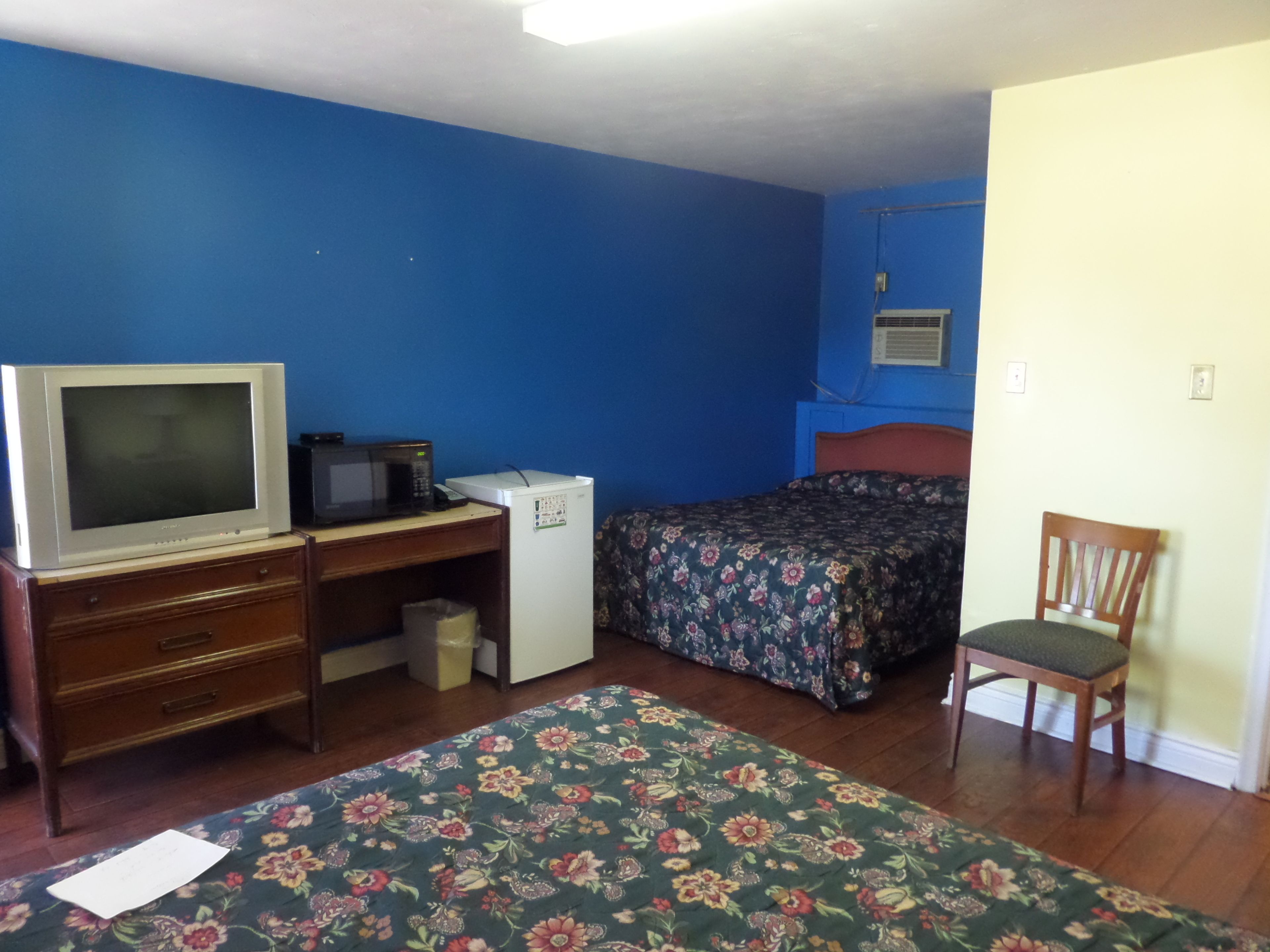 Quarto com cama, escrivaninha, televisão, frigorífico e uma cadeira.