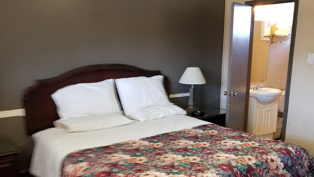 A hotel room with a large bed, a nightstand, a lamp, a bench, and a bathroom visible through an open door. - Lakeridge Motel (Whitby)