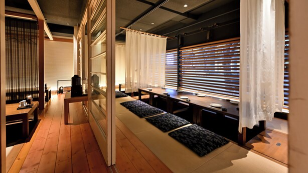 A modern interior with a long wooden bench, a dining table, and a view through large windows with blinds. - Richmond Hotel Hakataekimae (Fukuoka)