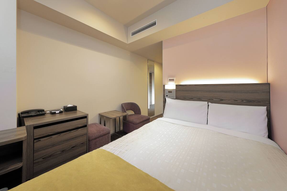 A hotel room with a bed, a desk with a chair, a small table, and a mirror.