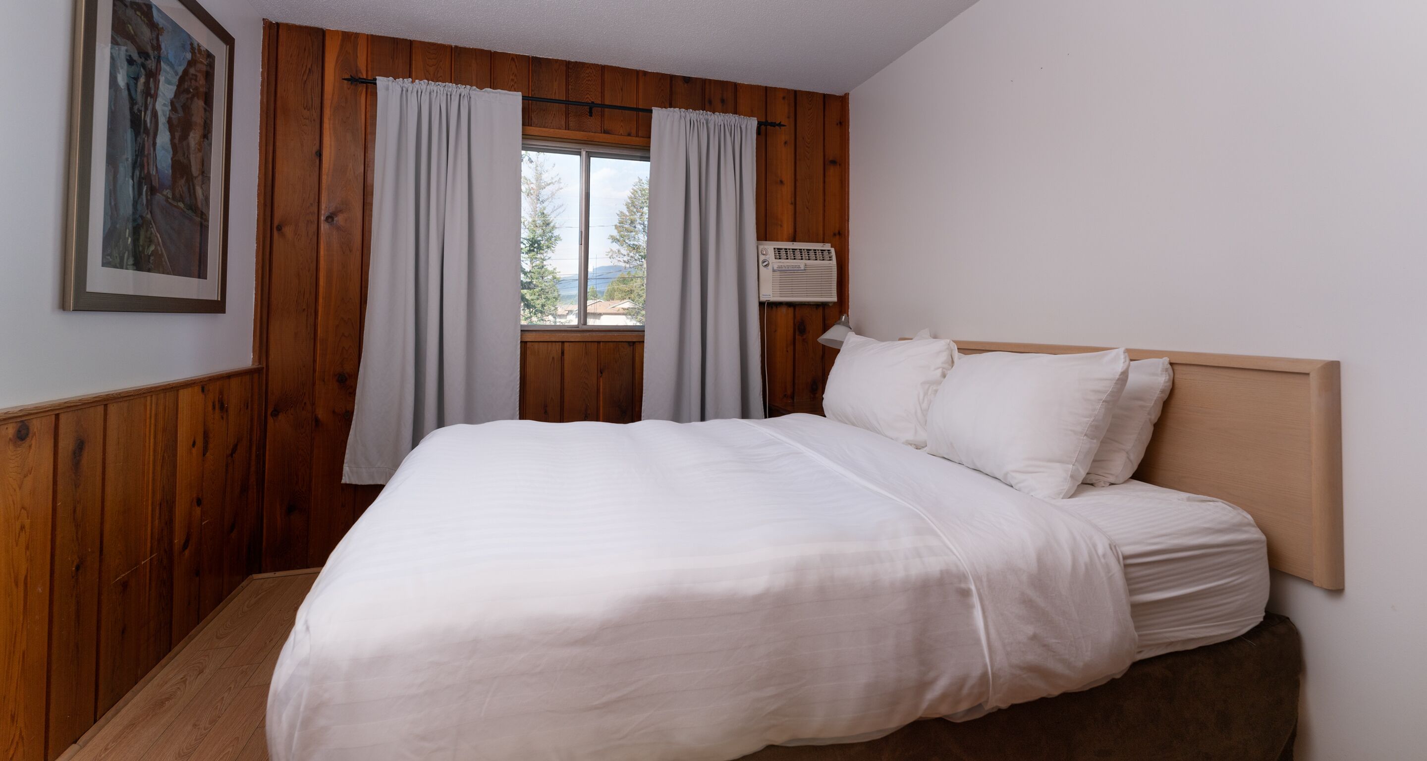 A bedroom with a wooden headboard, a large bed with white bedding, a window with curtains, and an air conditioning unit on the wall.