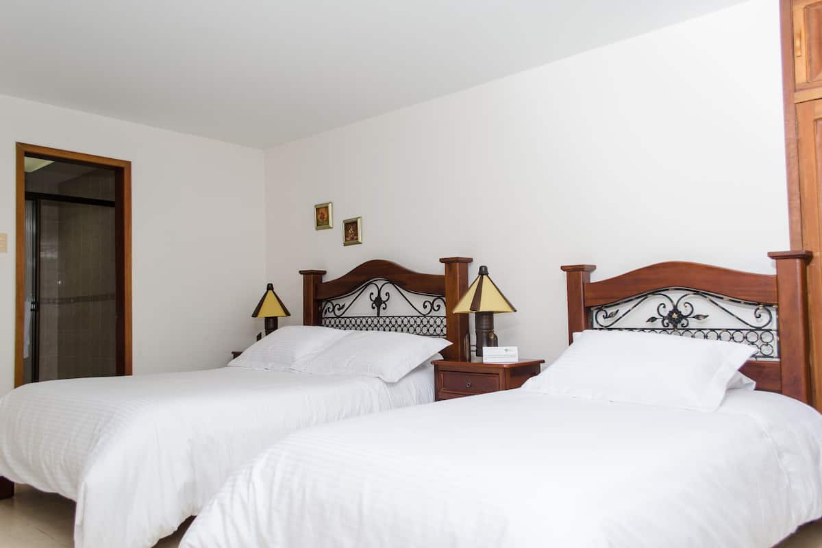 A hotel room with two single beds, wooden headboards, bedside tables with lamps, and a door leading to a bathroom.