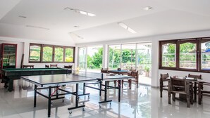 Amplio espacio interior con mesa de billar, equipo de billar y mesa de juego. Ventanas amplias ofrecen luz natural y vistas al exterior con vegetación.