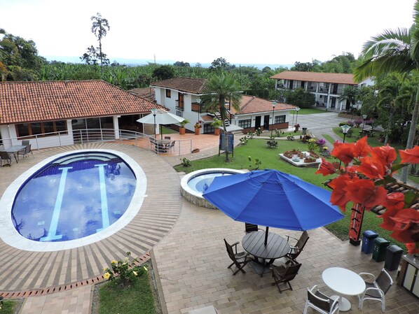 Un espace piscine comprenant une piscine circulaire, une plus petite piscine également circulaire, et une table avec des chaises placée sous un parasol bleu.