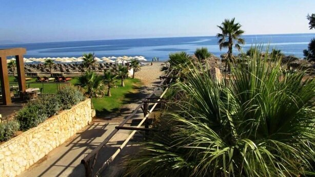 A coastal resort with a paved walkway leading to beach umbrellas and lounge chairs. - Panos Beach Hotel (Platanias)
