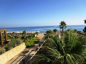A coastal resort with a paved walkway leading to beach umbrellas and lounge chairs. - Panos Beach Hotel (Platanias)