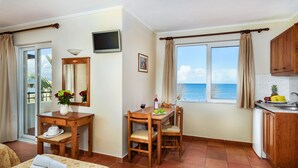 A room with a dining table, chairs, a TV, a window with curtains, a kitchen area, and a bed with towels. - Panos Beach Hotel (Platanias)