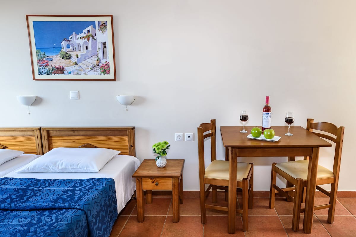 A hotel room with a bed, a small table with a vase, a dining table with chairs, and a painting of a coastal scene.