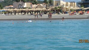 A beach with sun loungers and umbrellas, a boat, and buildings in the background. - Panos Beach Hotel (Platanias)