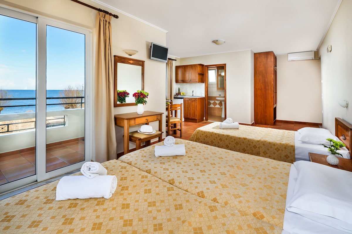 A hotel room with two beds, a balcony with a view of the sea, a kitchenette, and a TV.