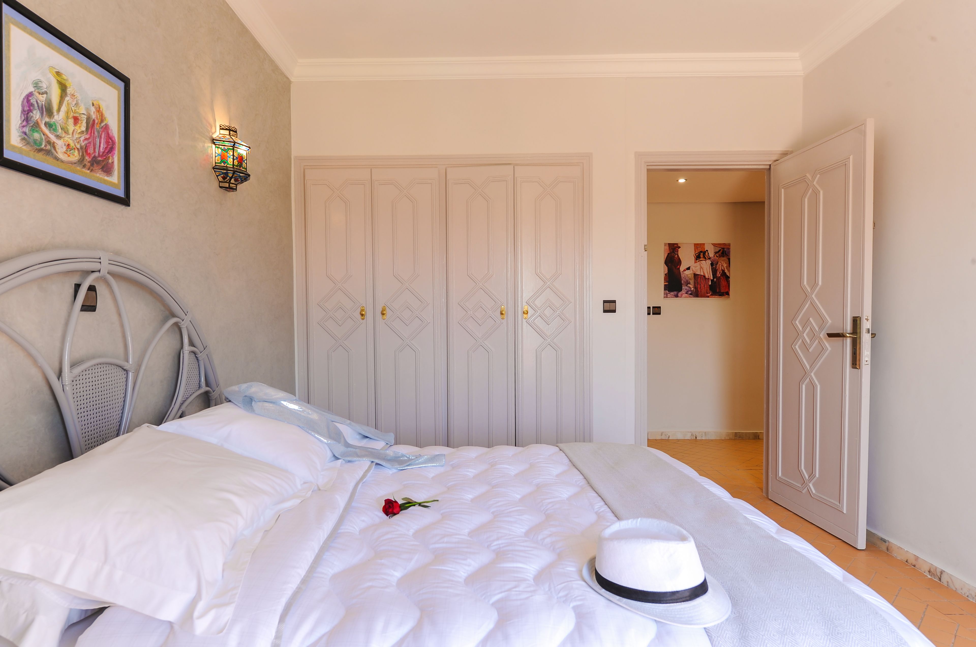 Una cama bien hecha con sábanas blancas, una sola rosa roja y un sombrero blanco sobre la cama. Hay una lámpara de pared y un cuadro enmarcado encima de la cama.