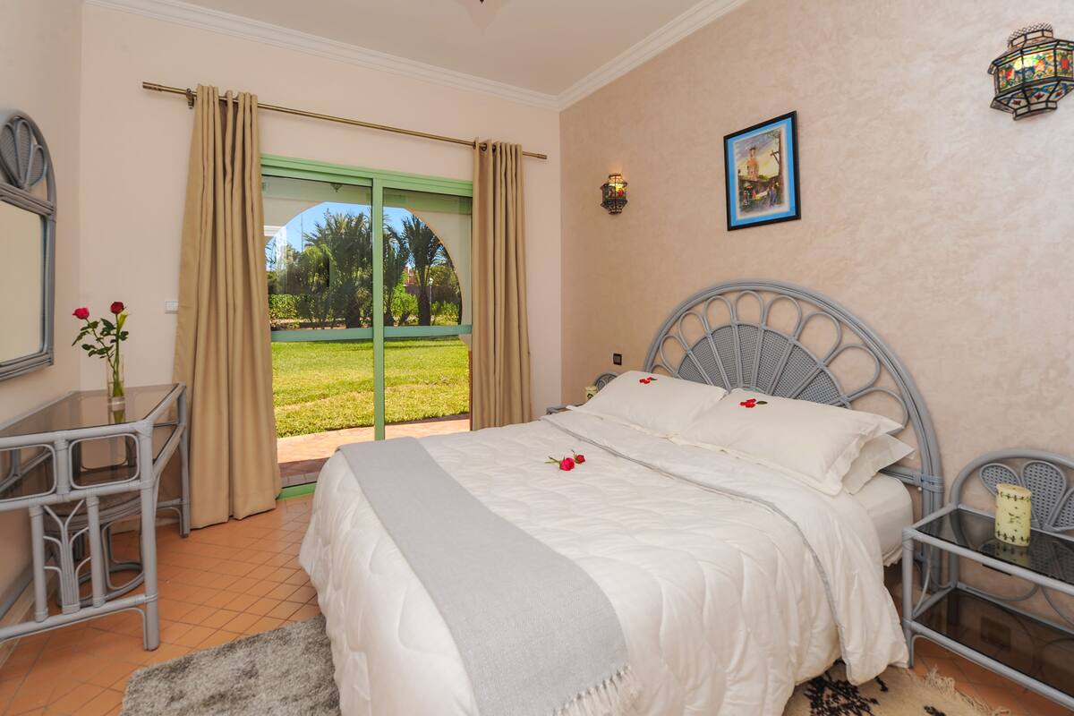 A bedroom with a bed, a desk, and a chair. There is a view of a garden through a sliding door.