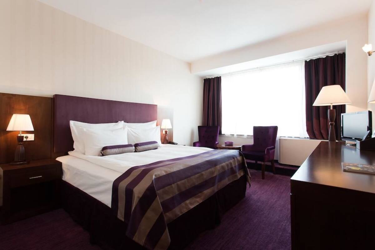 A hotel room with a large bed, two bedside lamps, a desk with a computer, a chair, and a window with curtains.