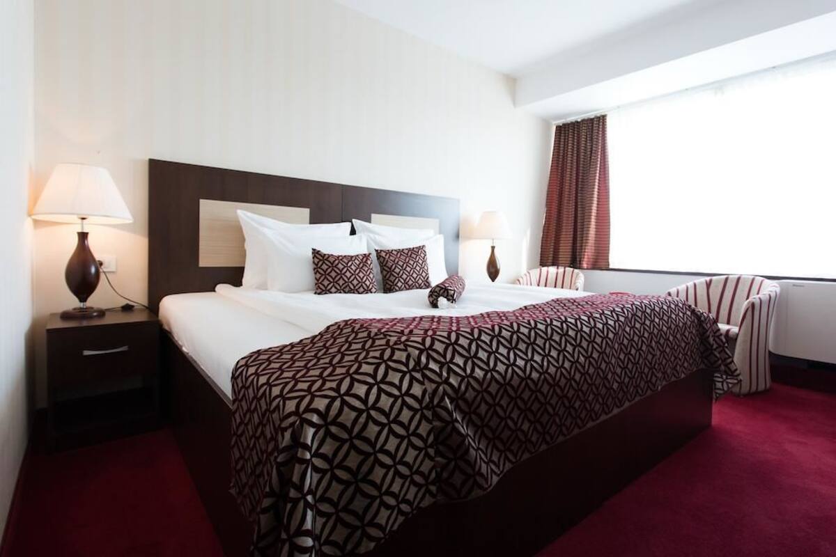A hotel room with a large bed, two bedside tables with lamps, a window with curtains, and a red carpet.