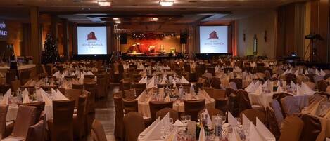 A banquet hall with round tables set for a formal event, a stage with a band, and a large screen displaying the hotel name.