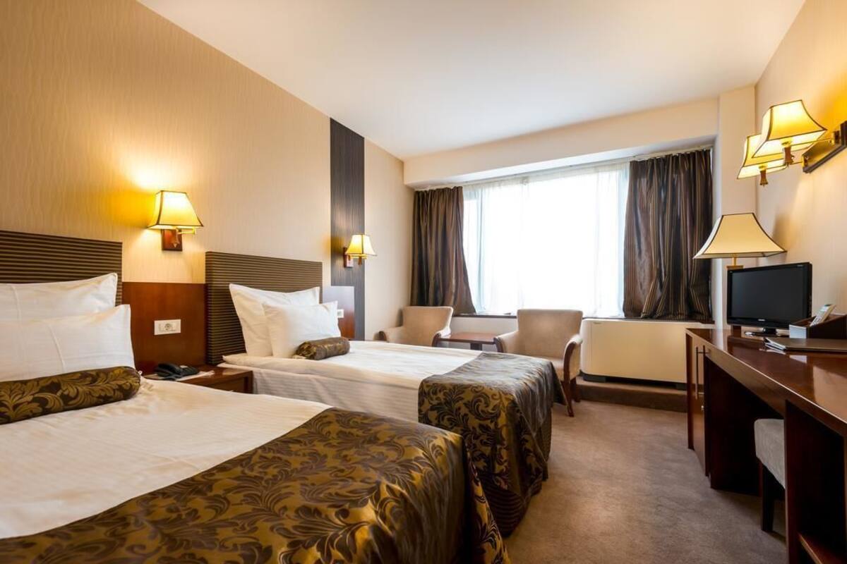 A hotel room with two beds, a desk, a chair, a TV, and a window with curtains.