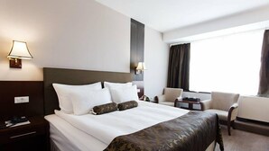 Une chambre d’hôtel avec un grand lit, deux tables de chevet, une chaise, un bureau et une fenêtre avec des rideaux.