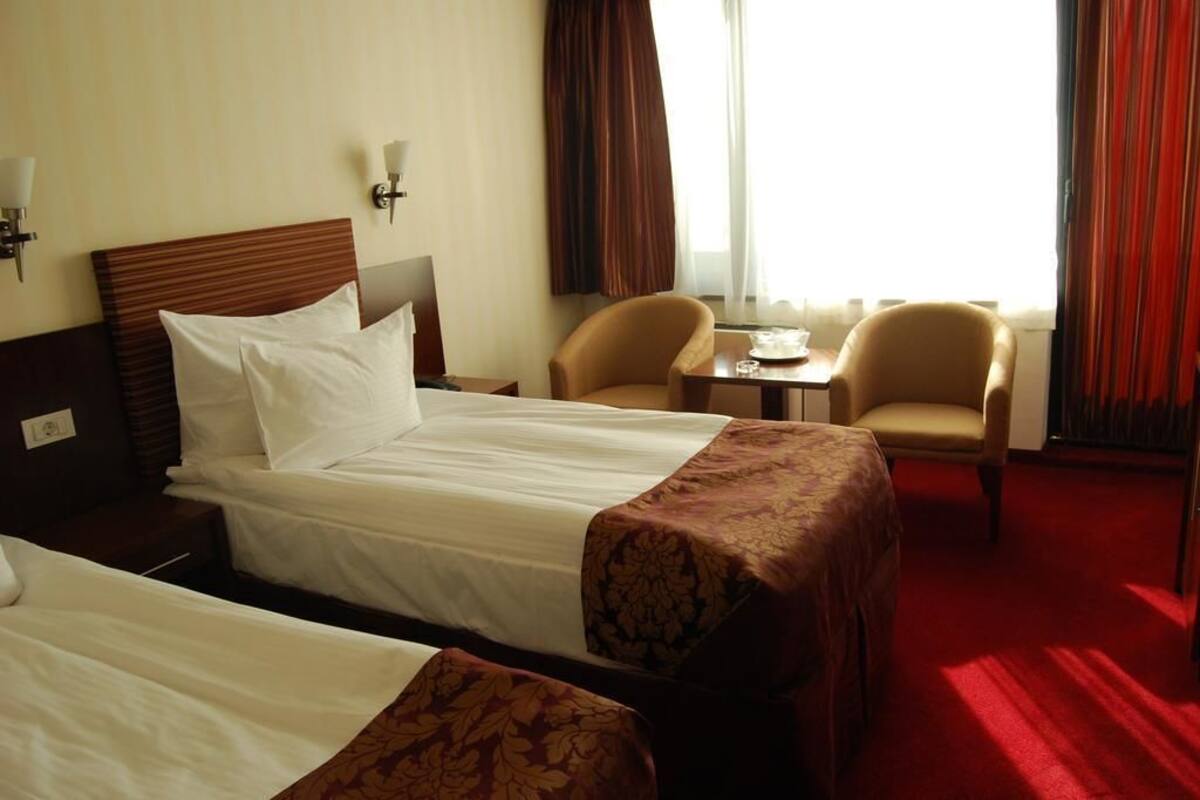 A hotel room with two beds, a small table, two armchairs, and a window with curtains.