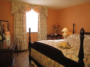 A bedroom with a bed, a bedside table, a lamp, a mirror, and a window with curtains. - Colston Hall Cottages (Woodbridge)