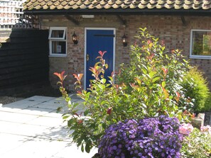A brick house with a blue door, a small window, and a garden with purple flowers. - Colston Hall Cottages (Woodbridge)