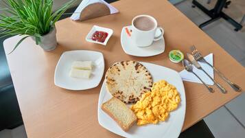 A breakfast setting with a plate of scrambled eggs, toast, a grilled flatbread, a cup of coffee, a small bowl of ketchup, and a plant on the table.
