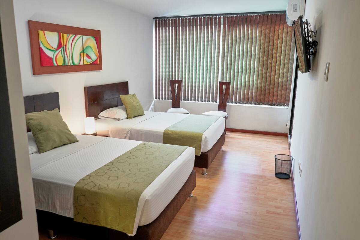 A hotel room with two beds, a large window with blinds, a painting on the wall, and a small trash bin.