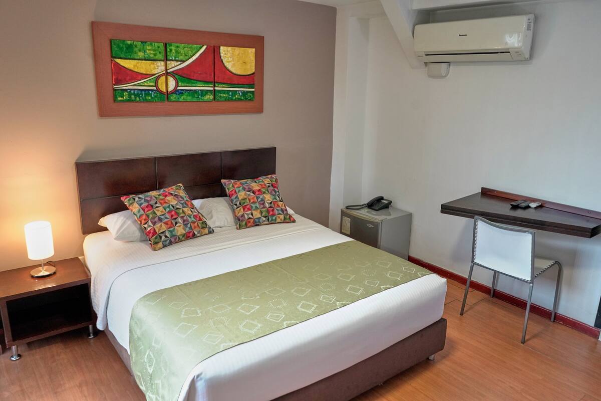 A hotel room with a bed, a nightstand with a lamp, a desk with a chair, and an air conditioner on the wall.