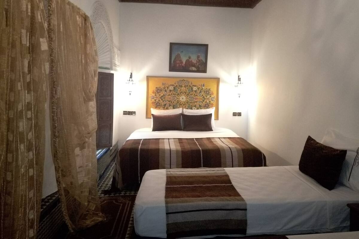 A hotel room with a bed, a headboard with a floral design, two bedside lamps, and a window with curtains.