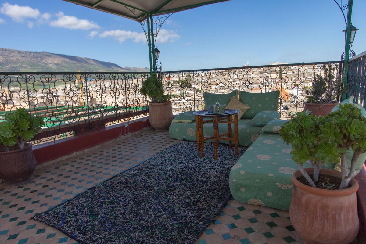 A terrace with a patterned floor, a green cushioned sofa, a small wooden table, and potted plants.