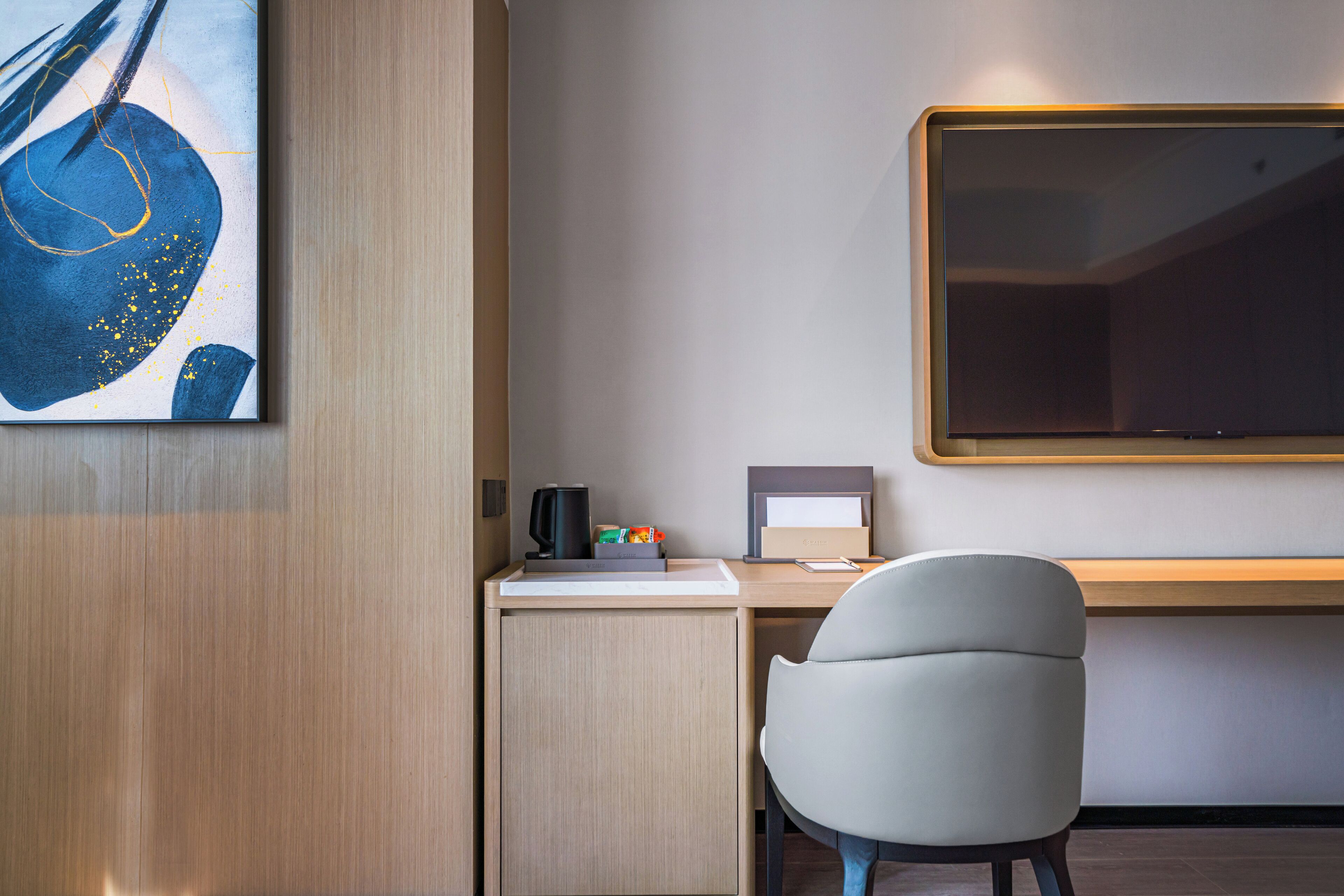 Quarto de hotel moderno com uma escrivaninha de madeira, uma cadeira branca, um quadro grande e uma TV de tela plana na parede.