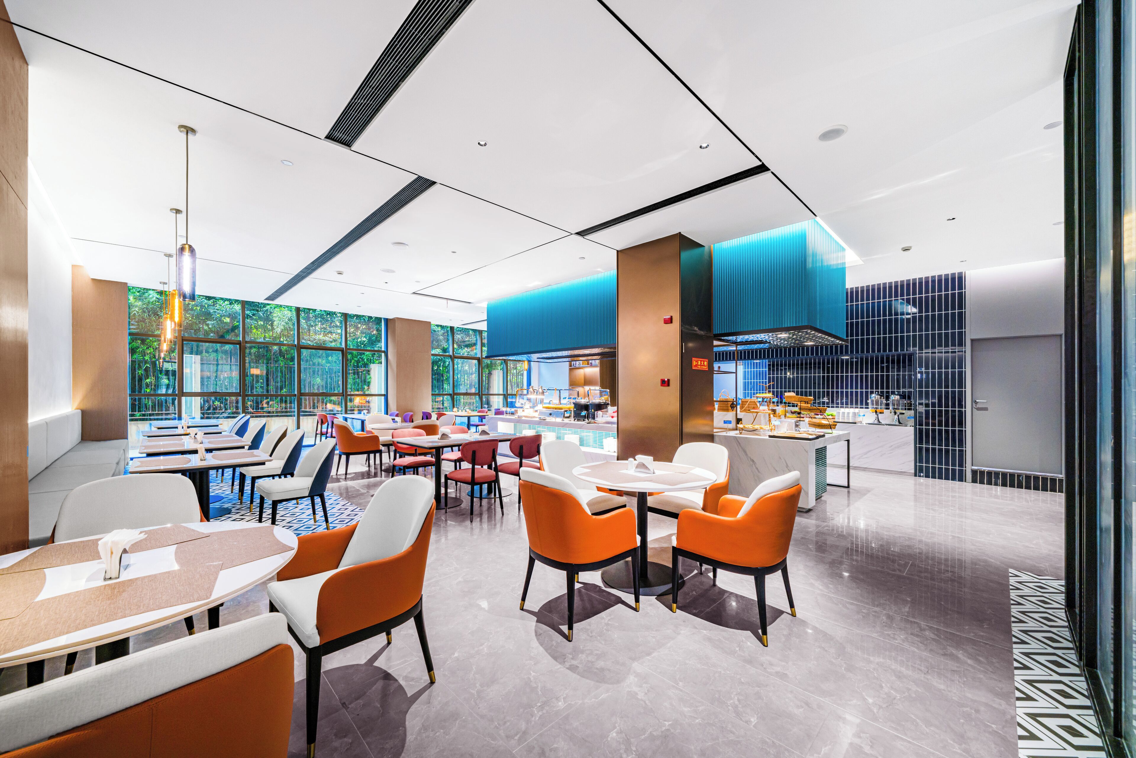 A modern dining area with tables and chairs, a kitchen in the background, and large windows allowing natural light.