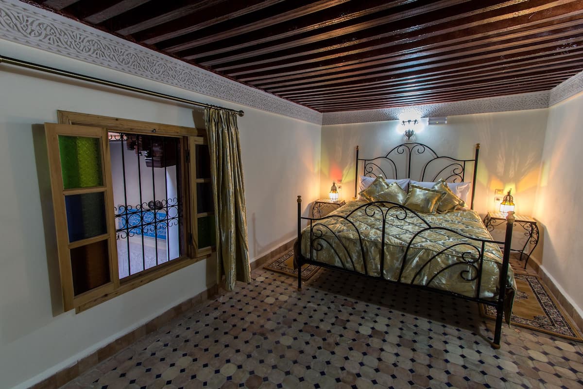 A bedroom with a bed, a window with curtains, a patterned floor, and a decorative ceiling.