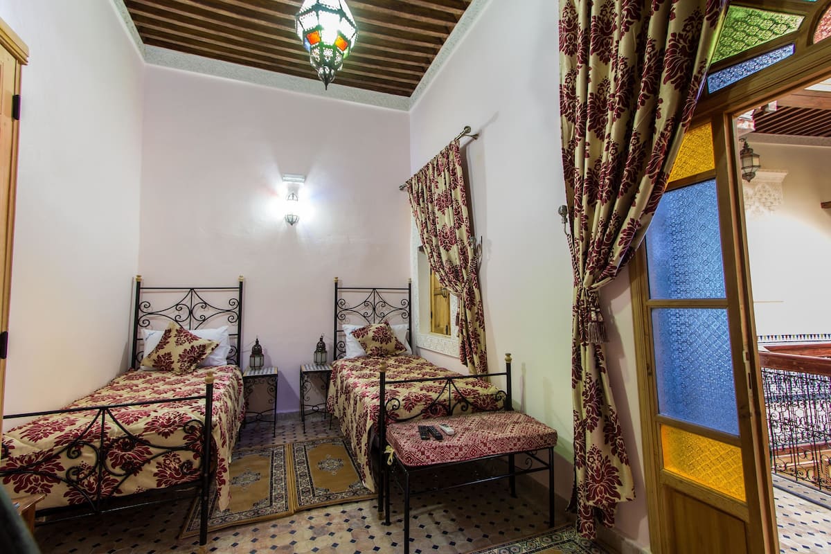 A room with two beds, a bench, a small table, and a window with patterned curtains.