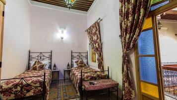 A room with two beds, a bench, a small table, and a window with patterned curtains.
