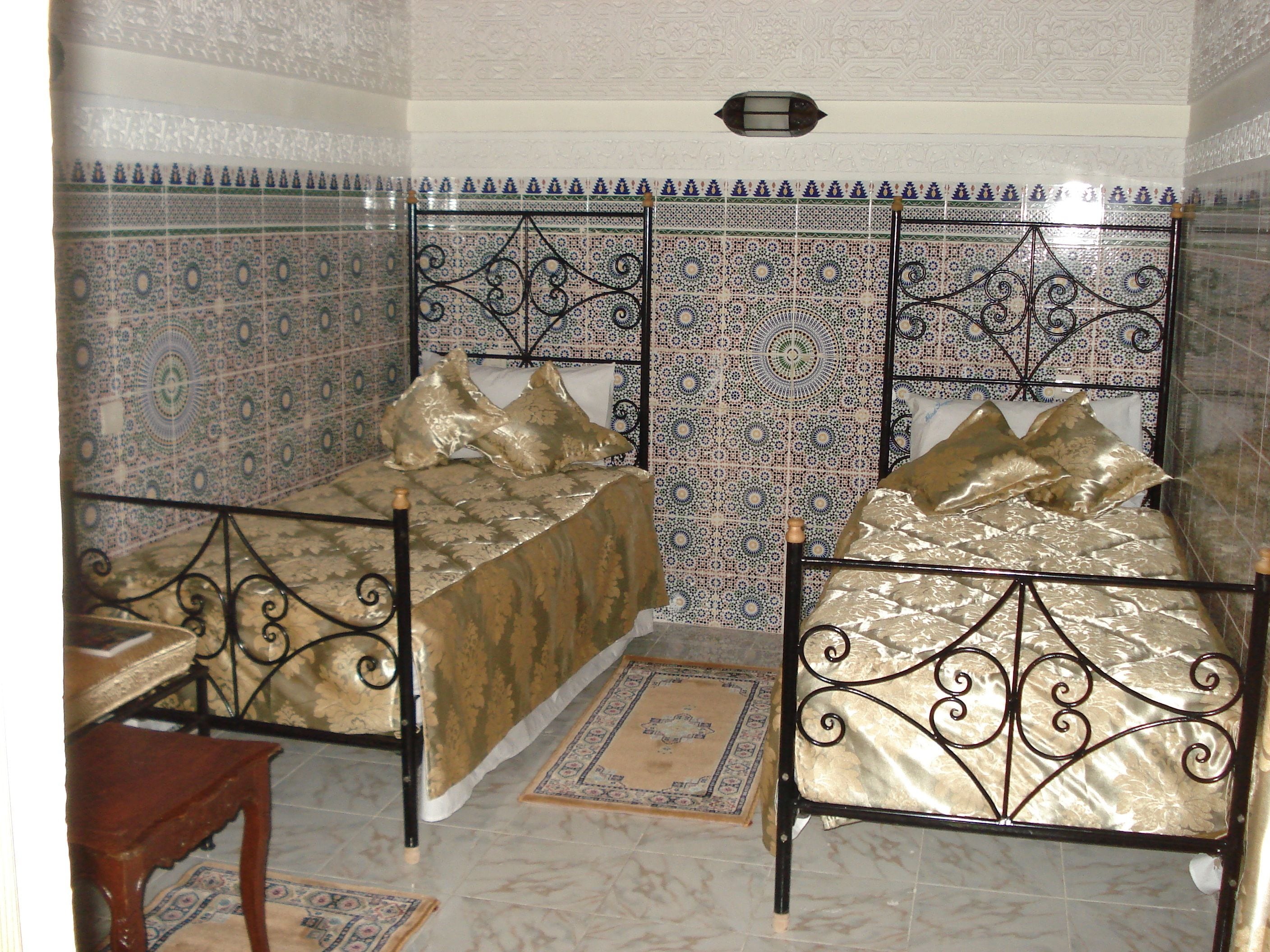 A room with two beds, each with a metal frame and a patterned bedspread. The walls are tiled with a blue and white geometric design.