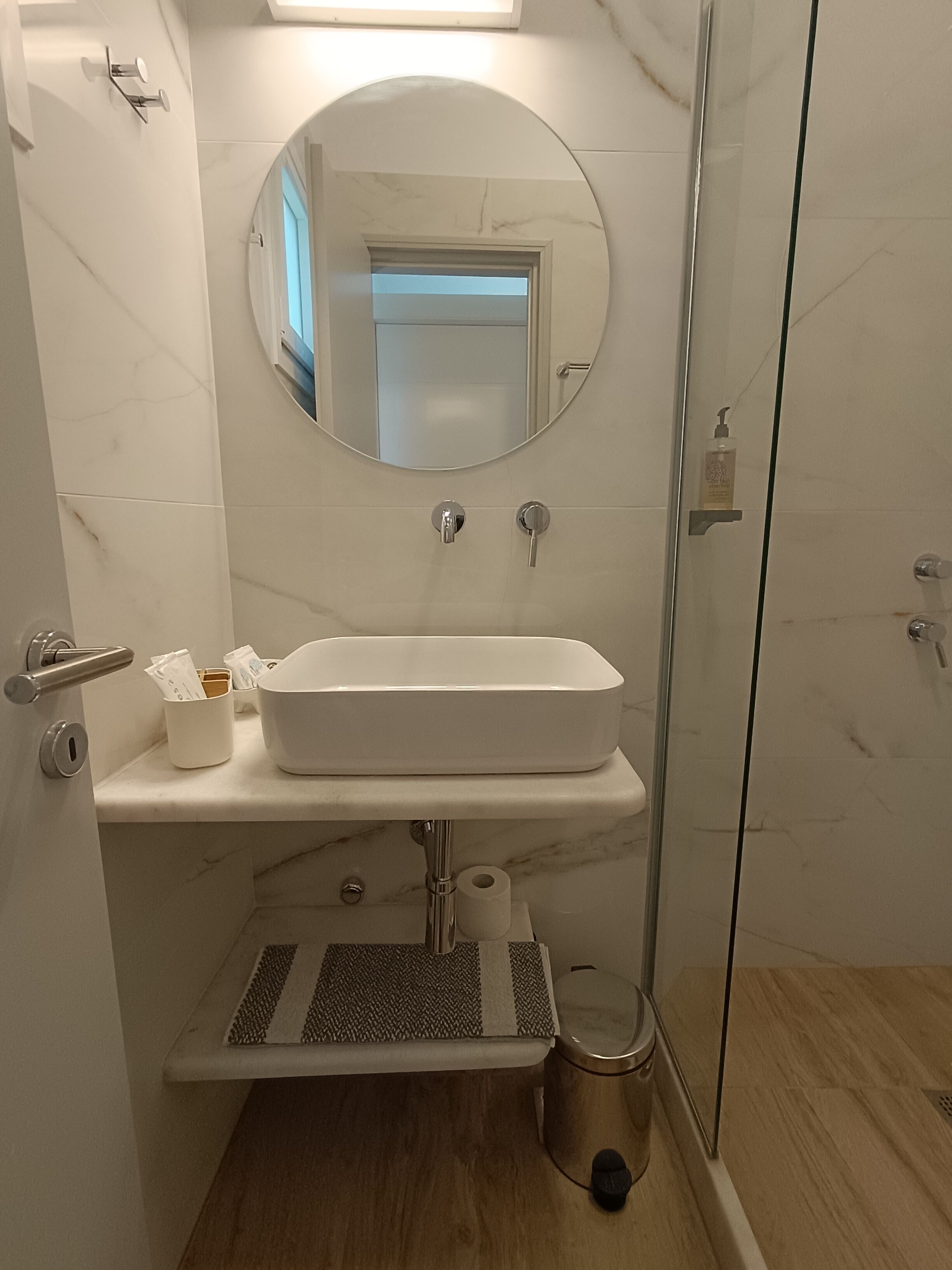 Egy fürdőszoba, amelyben fehér mosdókagyló, kerek tükör, zuhanyzó és WC található.