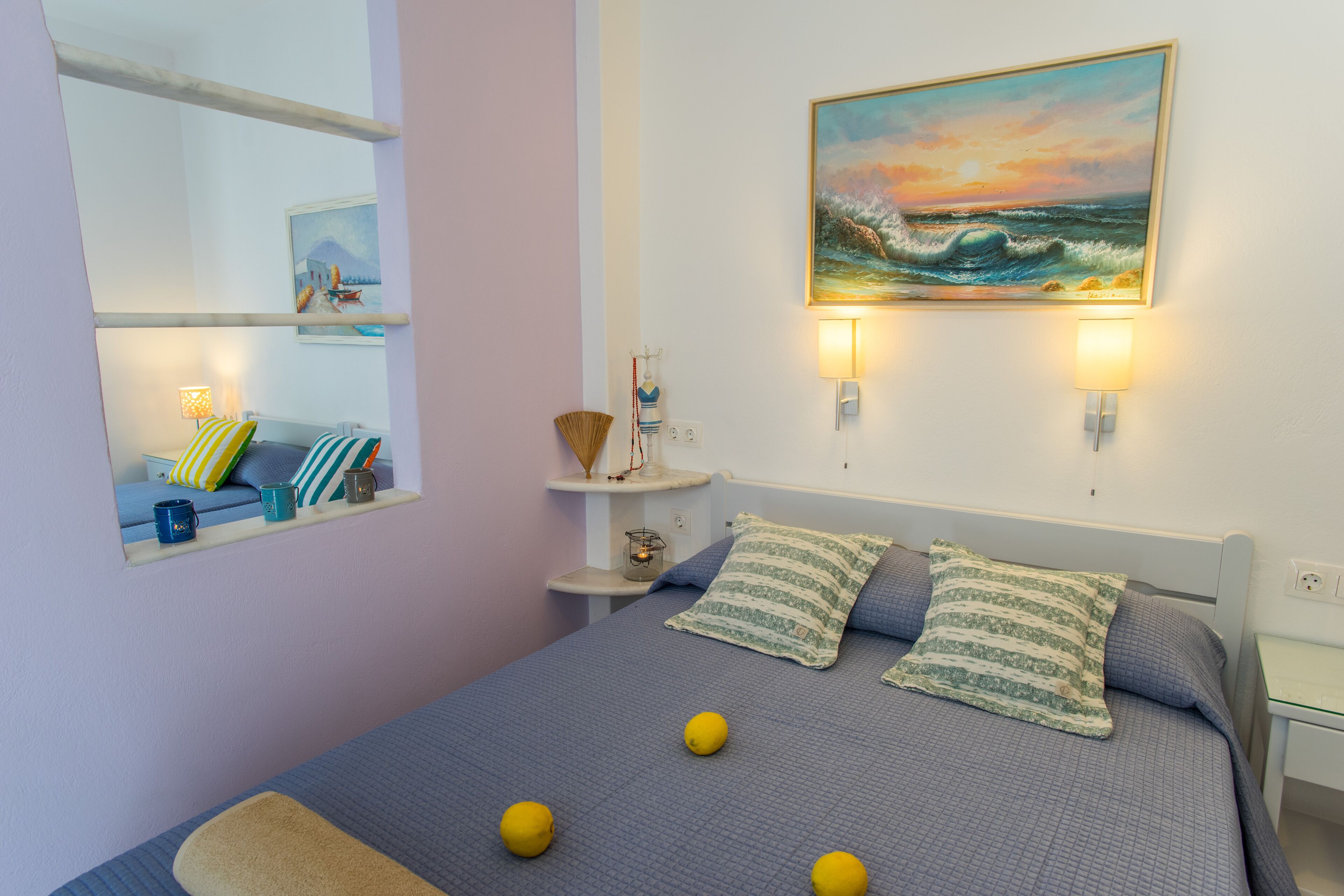 Une chambre à coucher comprenant un lit, des oreillers, une table de chevet, une lampe, une étagère avec des objets décoratifs et un tableau représentant un paysage marin.
