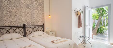 A bedroom with two beds, a nightstand, a hanging lamp, a chair, and a door leading to a garden.