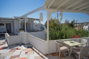 Uma área de estar coberta ao ar livre com mesa e cadeiras, vista para o mar e um edifício branco ao fundo.