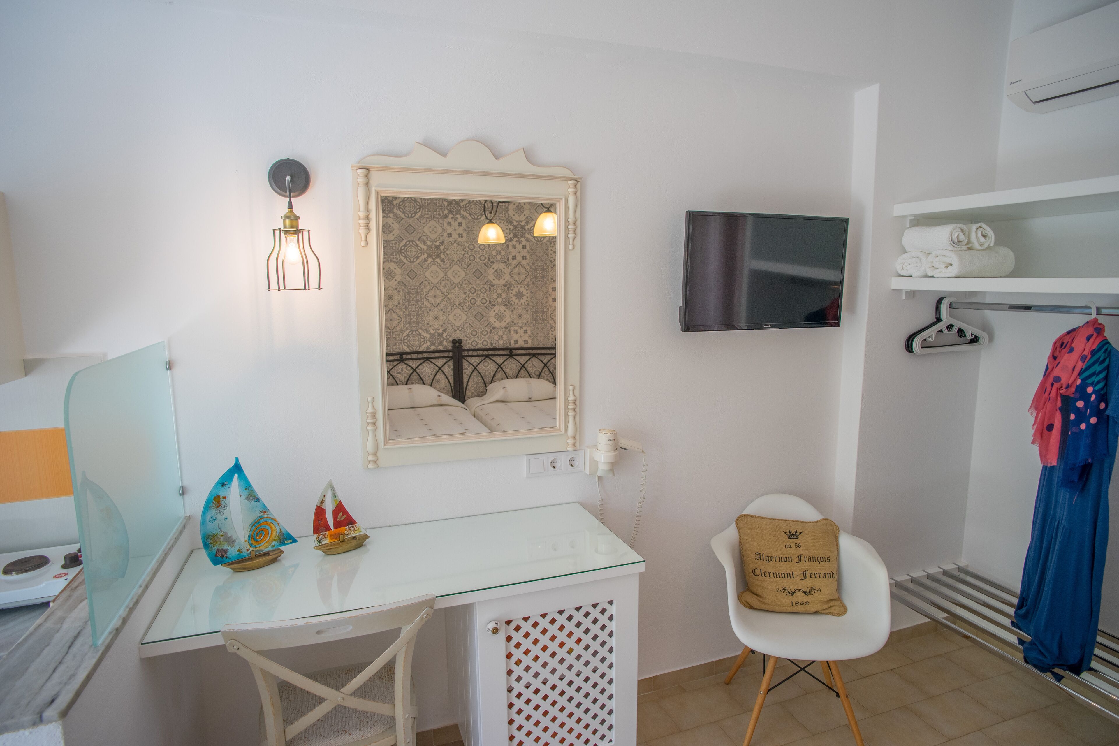 Habitació petita i ben il·luminada amb una taula de vidre, una cadira blanca, un llit amb capçalera estampada, una televisió i una estant amb tovalloles doblegades.